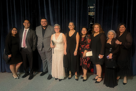 A group of nine adults poses in formal attire in front of dark blue stage curtains at what appears to be a gala or evening event. The group consists of both men and women dressed in elegant gowns, cocktail dresses, suits, and ties. Some individuals hold small clutches or purses, and all are smiling warmly. The ambiance suggests a celebratory or professional occasion, such as an awards night or community recognition event.