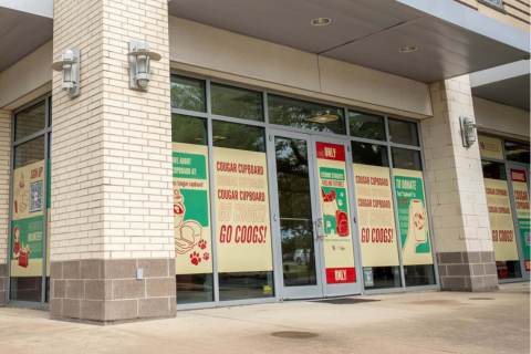 Front entrance of the Cougar Cupboard at the University of Houston, with window graphics promoting messages like ‘Feeding Students, Feeding Futures’ and encouraging campus support