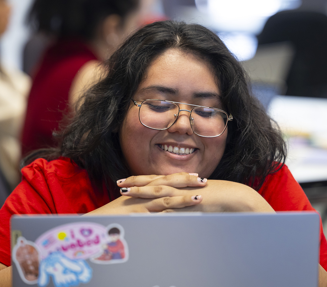 Student with brown hair and glasses, smiling looking at a laptop